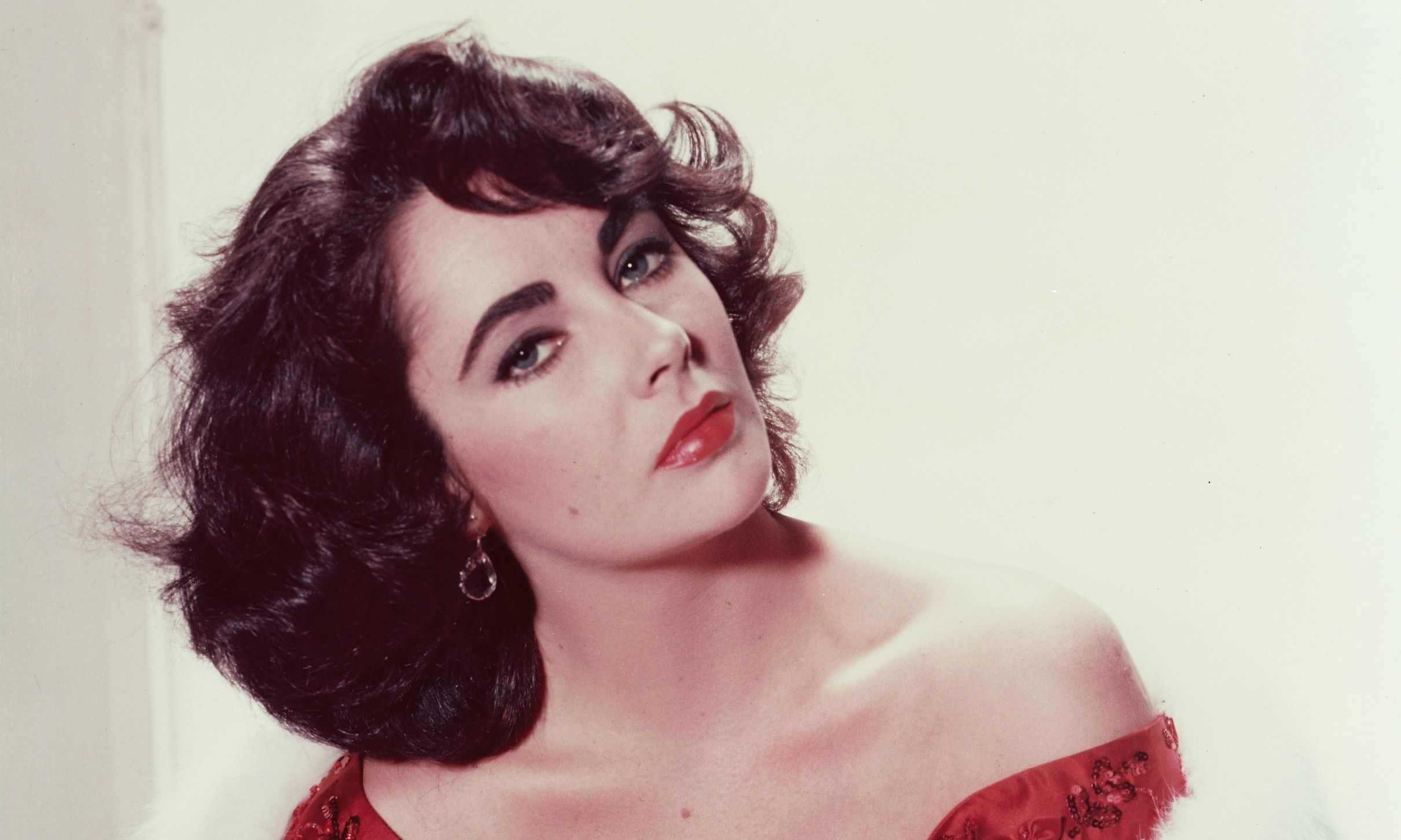 Elizabeth Taylor (1932-2011), British actress, wearing a red dress with a white fur stole in a studio portrait, against a white background, circa 1955.