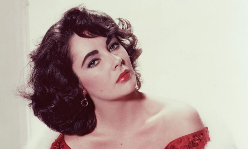 Elizabeth Taylor (1932-2011), British actress, wearing a red dress with a white fur stole in a studio portrait, against a white background, circa 1955.