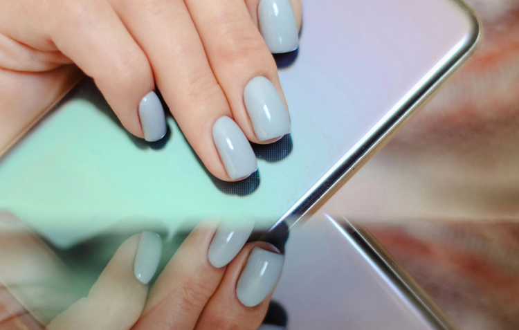 Close-up of a hand with glossy pale blue-gray gel manicure reflected in a mirrored phone case, nails resting against the device’s edge.