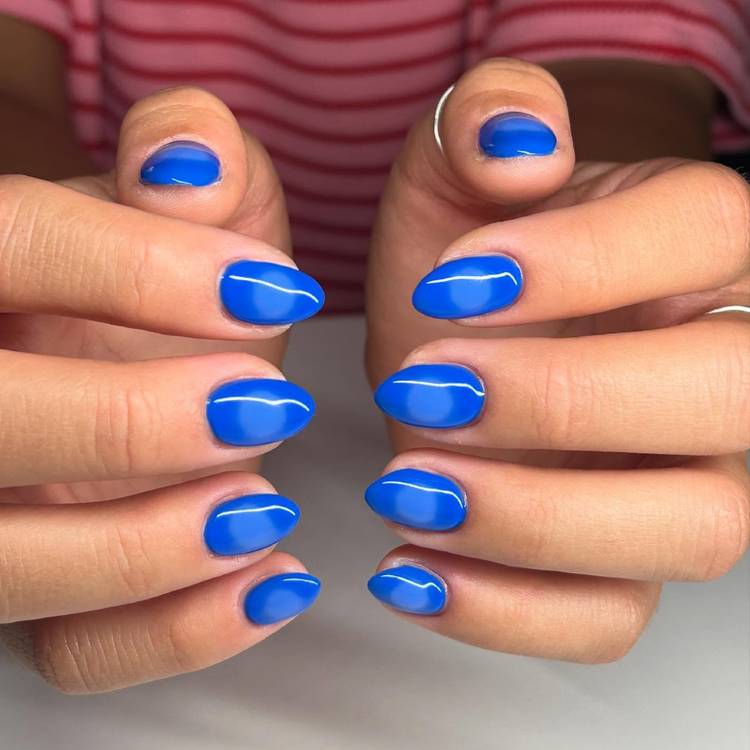 Close-up of two hands with short oval cobalt blue nails featuring a glossy halo nail effect with a lighter circular accent on each nail, photographed against a red and white striped top