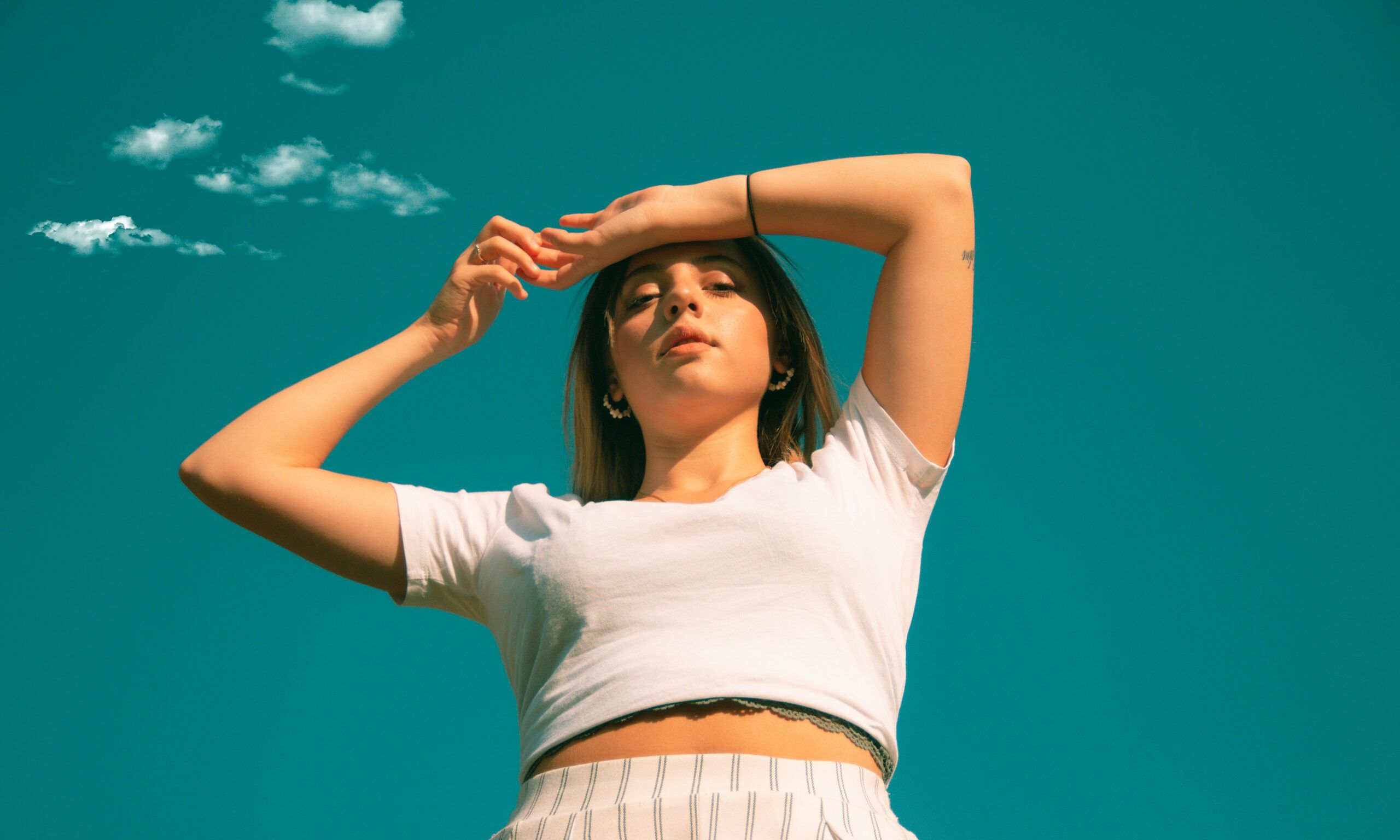 Up-angled photo of woman in white t-shirt with arms up by her forehead in front of teal background