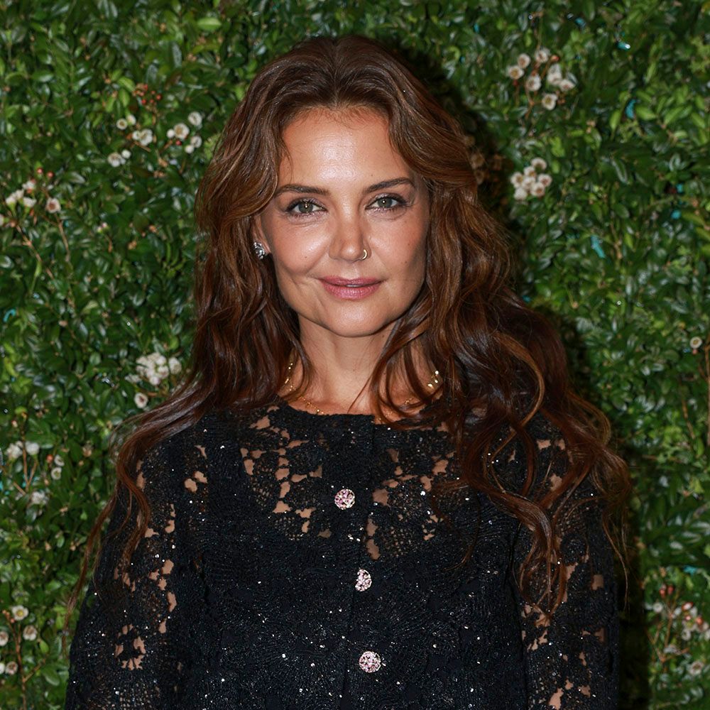 Katie Holmes wearing a black lace cardigan in front of a greenery shrub background