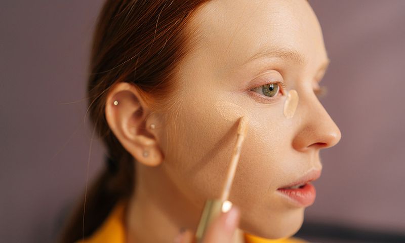 Close-up face of pretty young woman applying concealer on face under eye circles to correct skin in bedroom. Portrait of redhead female doing makeup putting base tone cream using foundation.