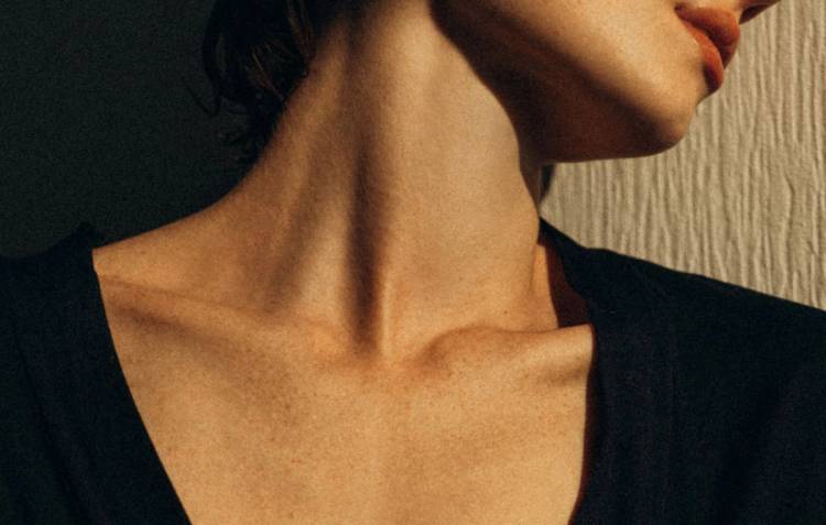 A close-up of a woman’s neck and décolleté in a black V-neck top, showing the chest area in warm light.