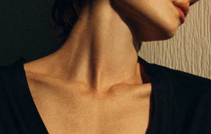 A close-up of a woman’s neck and décolleté in a black V-neck top, showing the chest area in warm light.