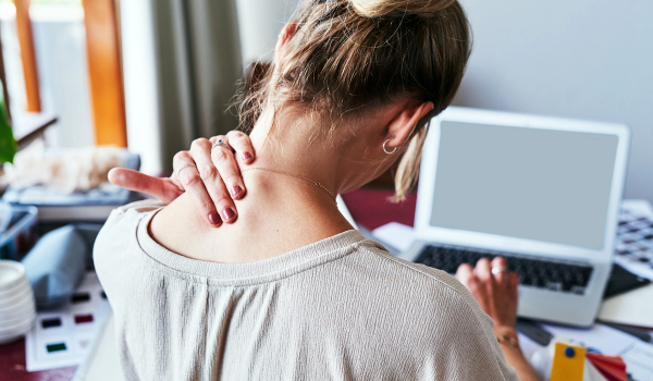 Woman seen from behind at a laptop, hand pressed to the back of her neck as she tilts her head forward.