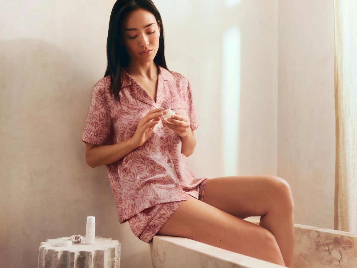 Model in Eberjey x ILIA pink silk pajama set applying ILIA skin care in a softly lit bedroom setting.