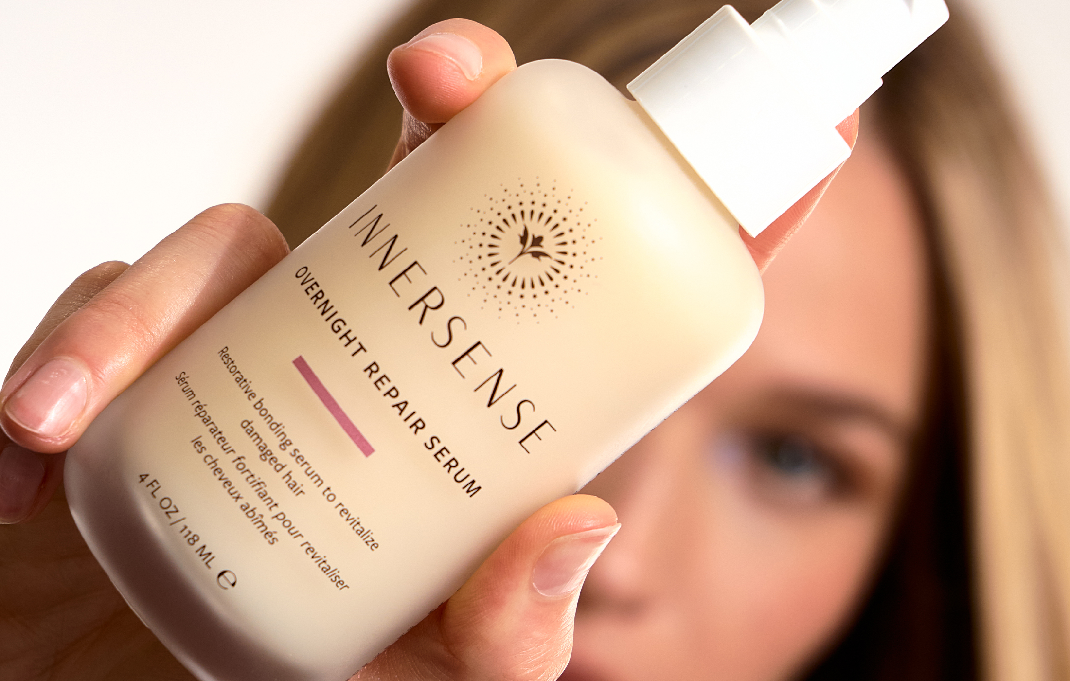 A woman holds a bottle of Innersense Overnight Repair Serum close to the camera, with her face softly blurred in the background.
