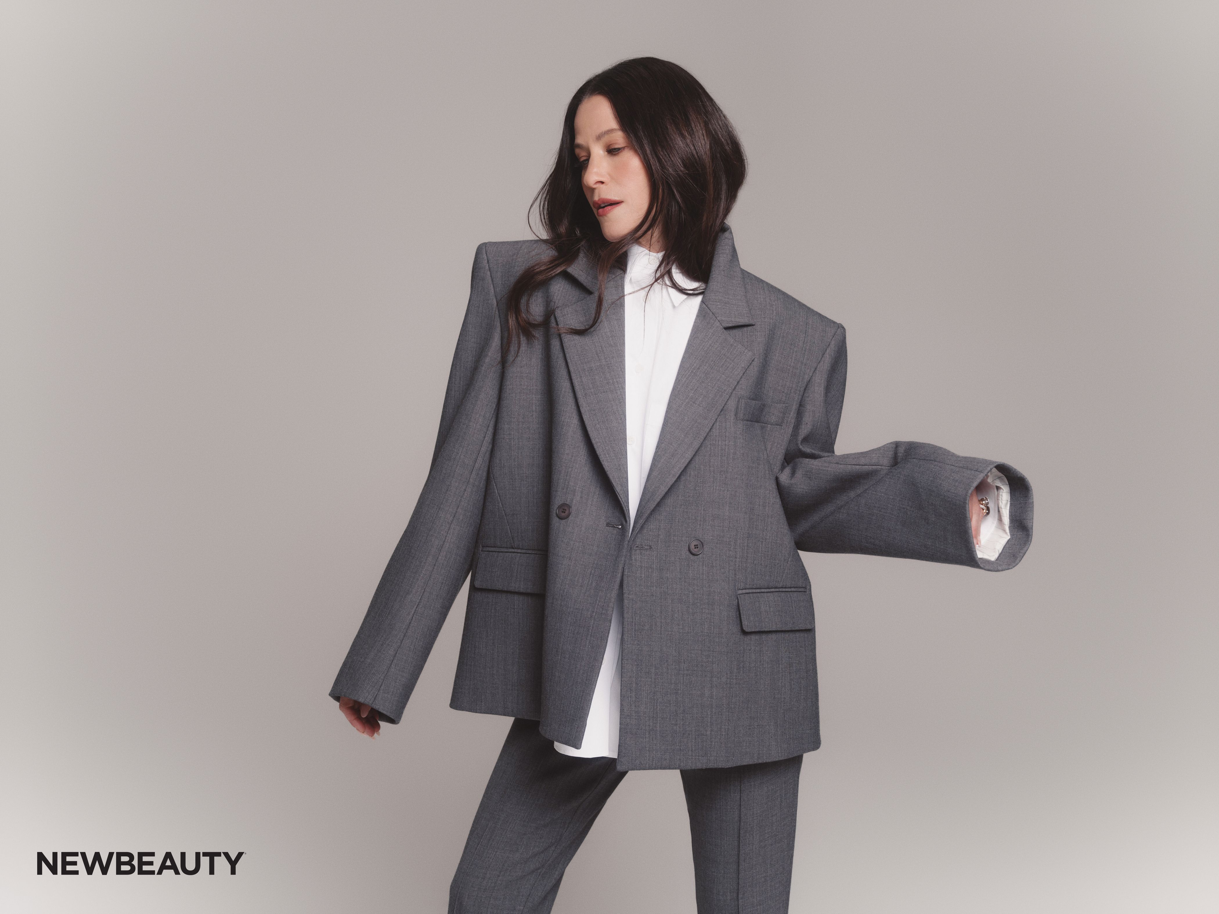 Jackie Tohn in an oversized gray suit and white shirt, posed mid-movement with one arm outstretched against a soft gray background.