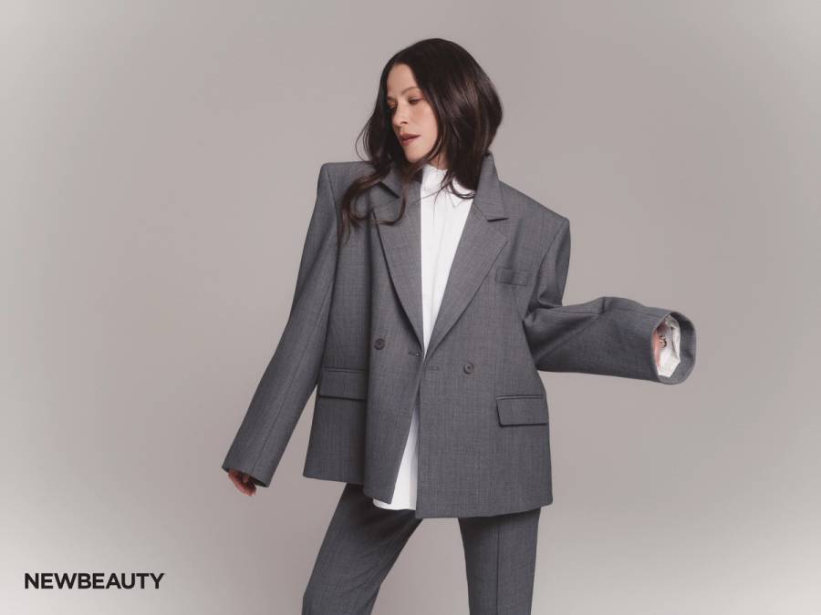 Jackie Tohn in an oversized gray suit and white shirt, posed mid-movement with one arm outstretched against a soft gray background.