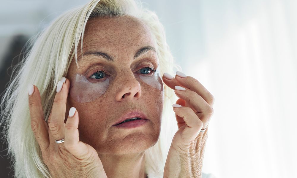 Older woman applying on her face hydrogel under-eye recovery patches. Do morning routine, to reduce puffiness. Skincare treatments usage, beauty routine concept