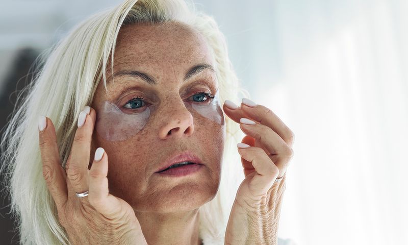 Older woman applying on her face hydrogel under-eye recovery patches. Do morning routine, to reduce puffiness. Skincare treatments usage, beauty routine concept