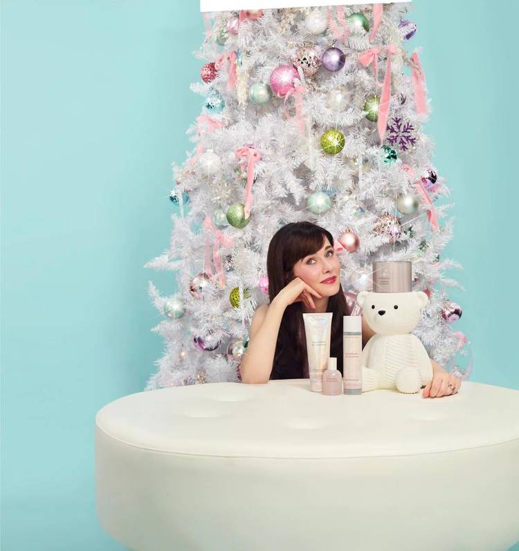 Zooey Deschanel rests her chin on her hand beside Bath & Body Works Snowflakes & Cashmere products and a white knit teddy bear