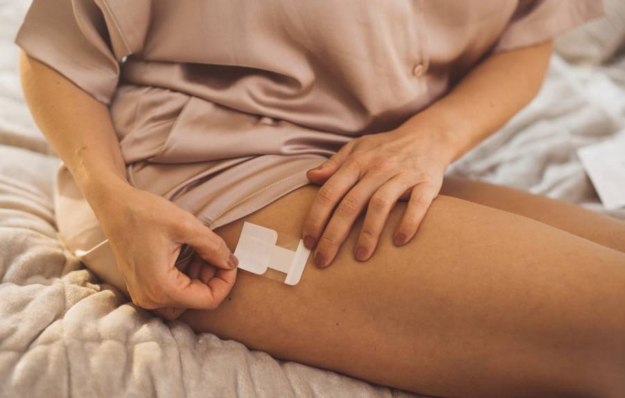 Close-up of a woman applying an estrogen patch to her upper thigh while sitting on a bed
