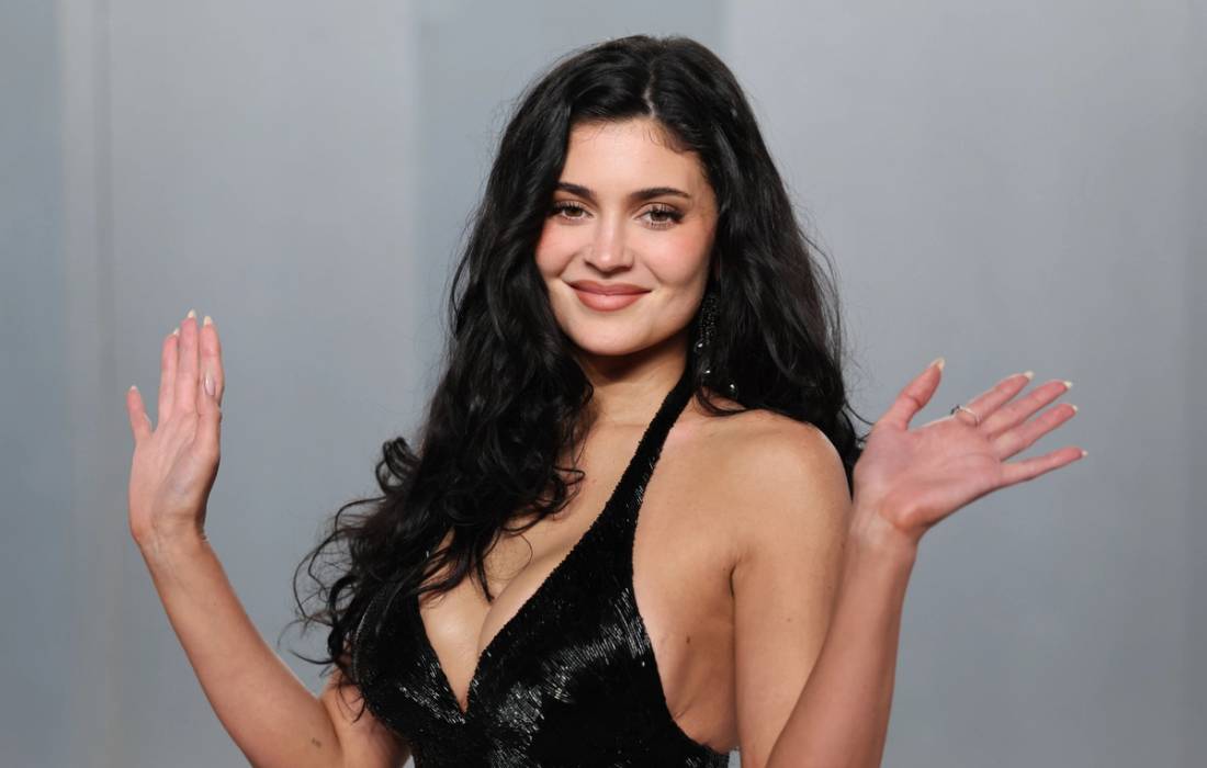 Kylie Jenner smiles and raises both hands while wearing a black sequined halter dress against a gray backdrop.
