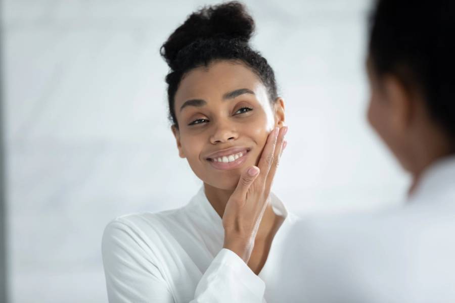 Smiling woman in a white robe touches her cheek while looking at her reflection in a bathroom mirror