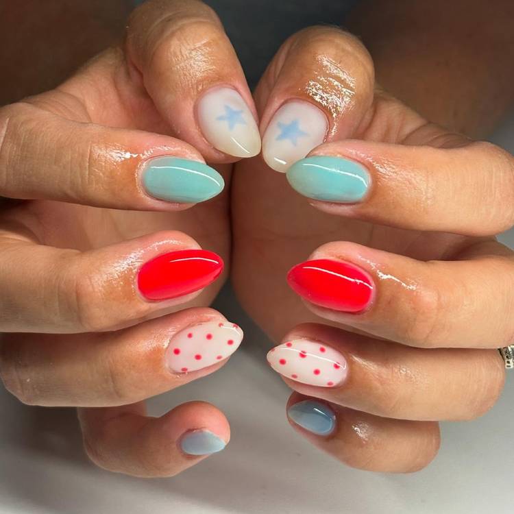 Close-up of two hands with a mixed pattern manicure featuring white nails with blue star accents, solid mint blue nails, bright red nails and white nails with red polka dots on almond-shaped nails