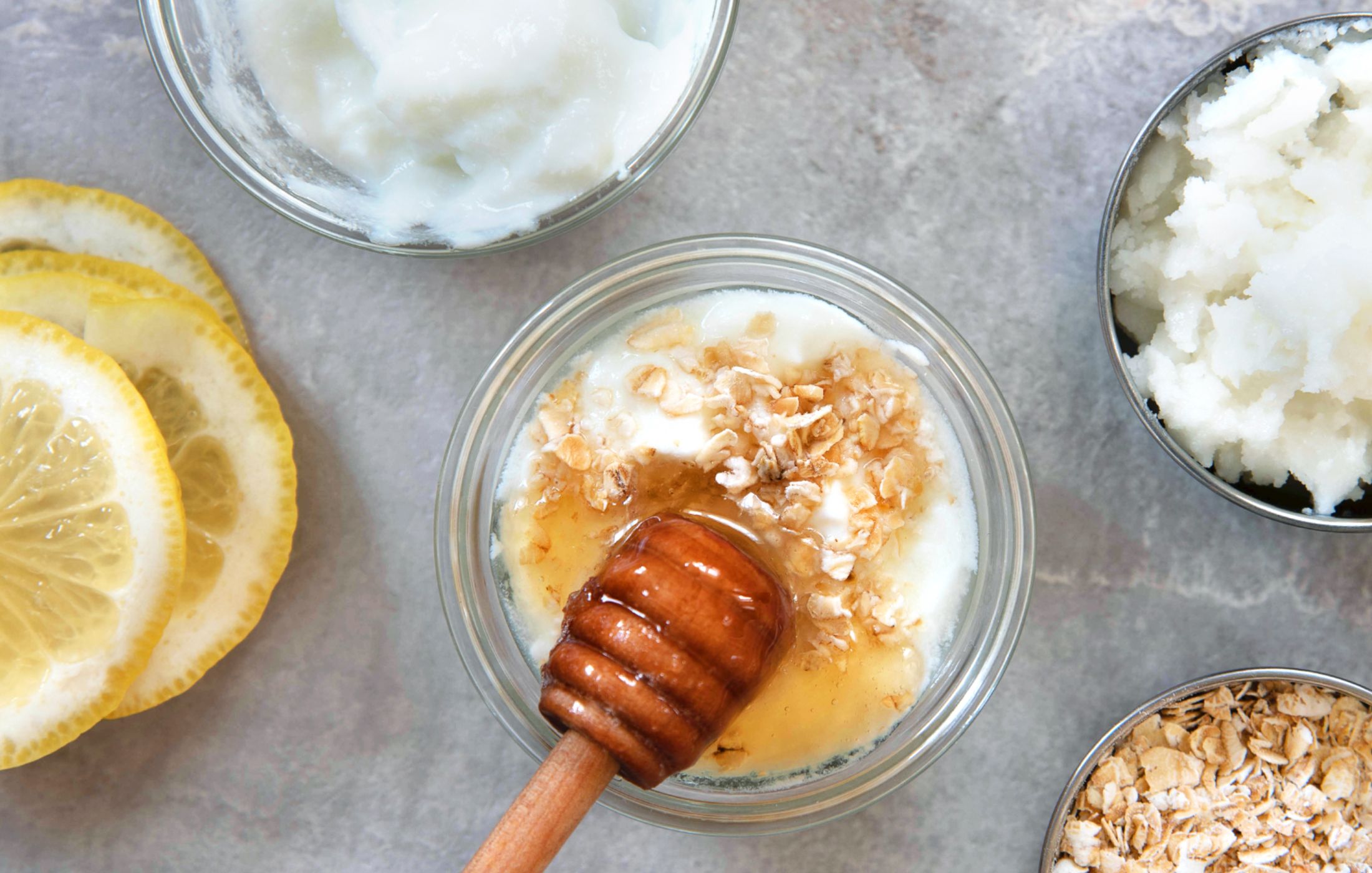 Overhead view of DIY face mask ingredients in small glass and metal bowls—plain yogurt, honey with a wooden dipper, rolled oats, a sugar scrub and sliced lemon—on a gray stone surface