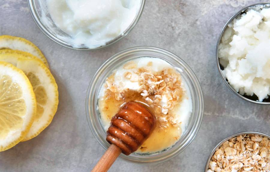 Overhead view of DIY face mask ingredients in small glass and metal bowls—plain yogurt, honey with a wooden dipper, rolled oats, a sugar scrub and sliced lemon—on a gray stone surface