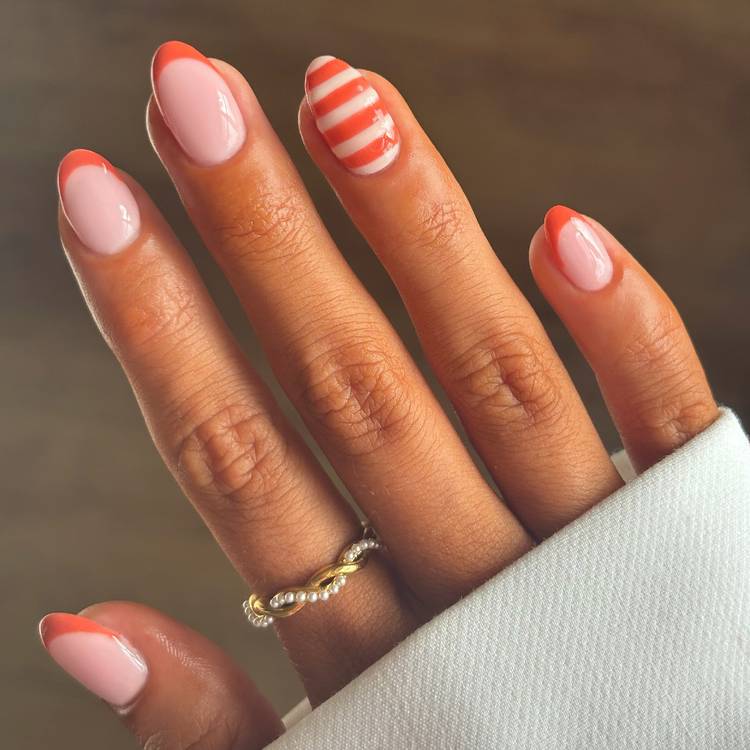 Short, rounded nails with a sheer pink base and bright orange tips. One accent nail has orange and white diagonal stripes.