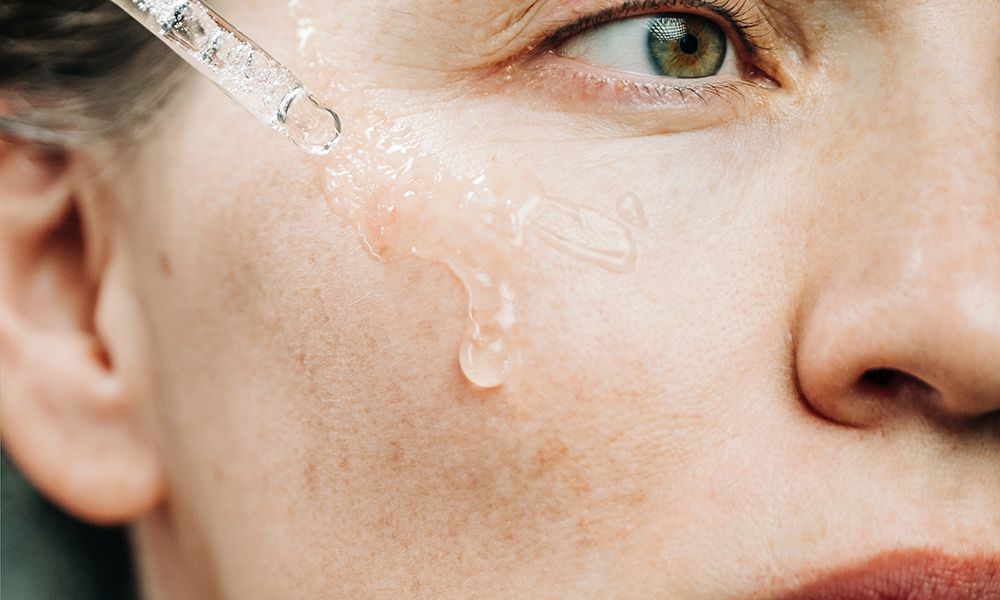 A beautiful woman delicately applies a serum to her cheek using a dropper in a dark, atmospheric setting. This intimate close-up captures self-care, natural beauty routine, and focus on skin health.