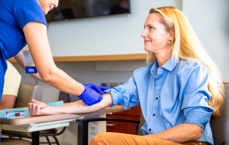 A nurse in blue scrubs prepares to draw blood from a smiling woman's arm during a clinic visit.