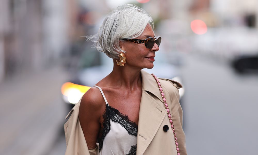 COPENHAGEN, DENMARK - AUGUST 07: Grece Ghanem is seen wearing large gold hoop earrings; oval sunglasses with a tortoiseshell frame from Prada; a beige trench coat; underneath a cream-white maxi dress with a V-neck, spaghetti straps, and black lace details from Gestuz; and a pink, blue and white patterned tweed bag from Chanel before the Gestuz Show during the Copenhagen Fashion Week (CPHFW) SS25 on August 07, 2024 in Copenhagen, Denmark.