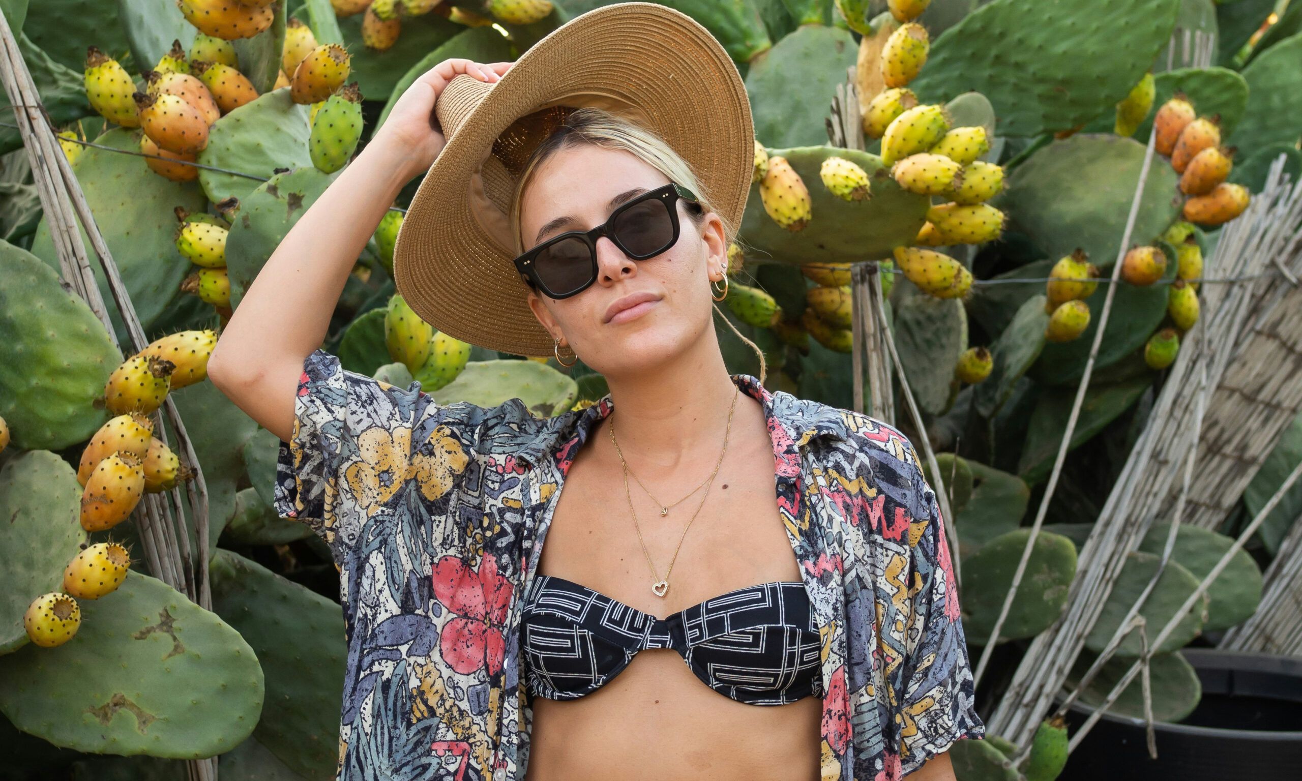 woman outdoors in front of cacti with bikini top, hat and sunglasses