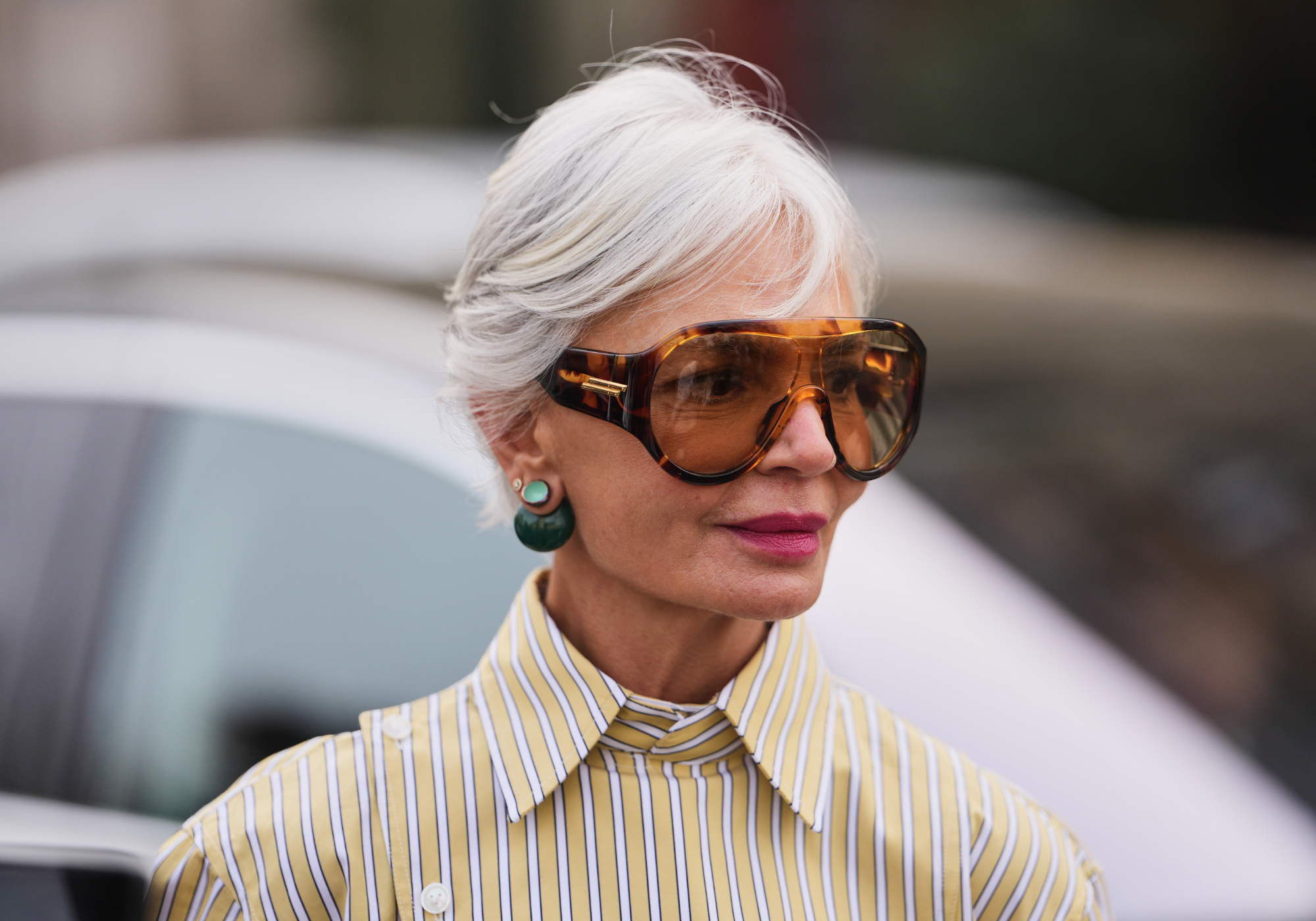 A woman with short silver hair wearing oversized tortoiseshell sunglasses, green drop earrings and a yellow striped shirt.