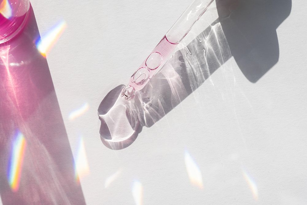Glass pipette with pink beauty serum on a white background with rainbow highlights