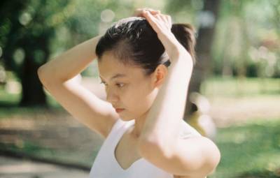 Woman pulling her hair into a ponytail outdoors, part of her scalp visible along the hairline.