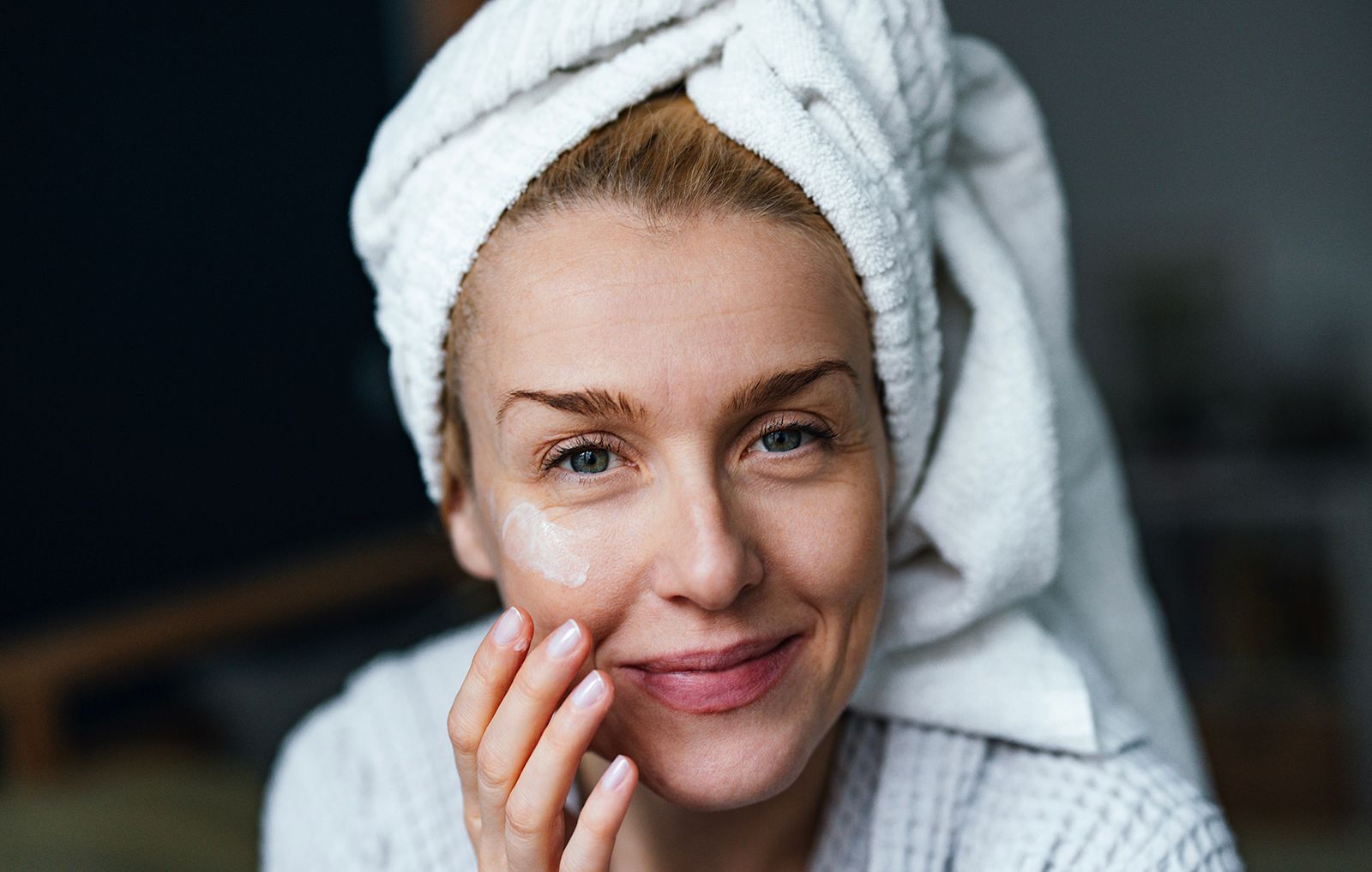 Close up photo of happy woman in bathrobe using face cream and looking at camera 