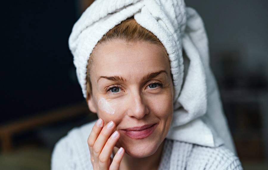 Close up photo of happy woman in bathrobe using face cream and looking at camera