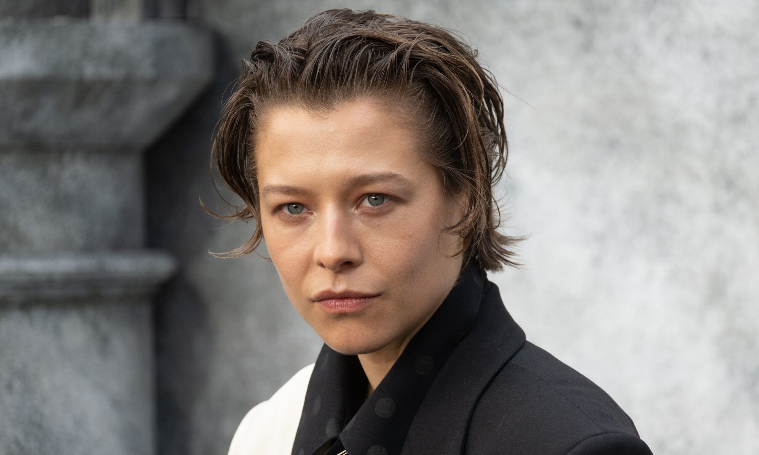 Emma D'Arcy in a black and white suit in front of a stone background