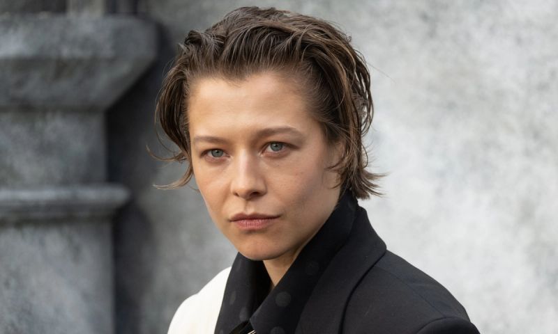 Emma D'Arcy in a black and white suit in front of a stone background