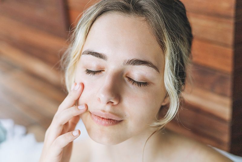 A young woman with blonde hair gently touches her face, her eyes closed. She appears to be in a relaxed state, possibly applying skincare.