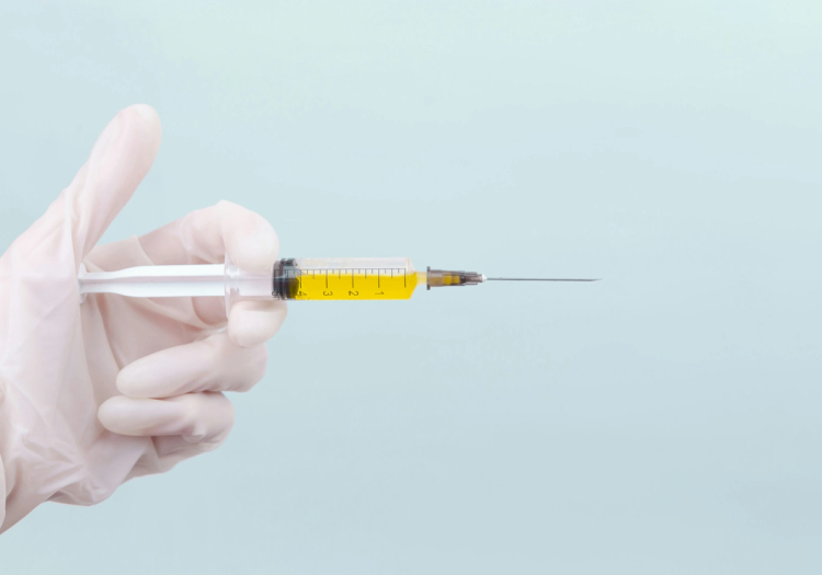 Gloved hand holding a syringe filled with yellow liquid against a pale blue background.