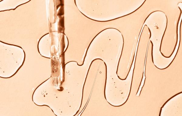 Close-up of a glass dropper releasing a serum, with squiggly lines of clear liquid on a peach-toned background