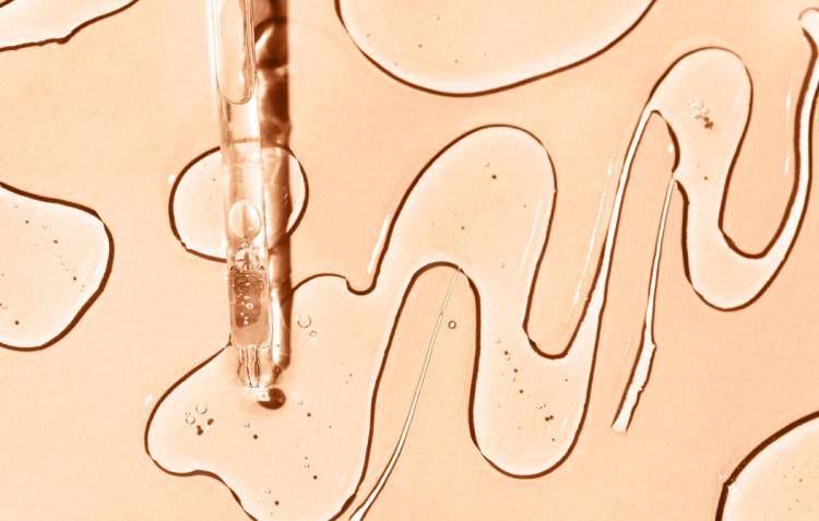 Close-up of a glass dropper releasing a serum, with squiggly lines of clear liquid on a peach-toned background