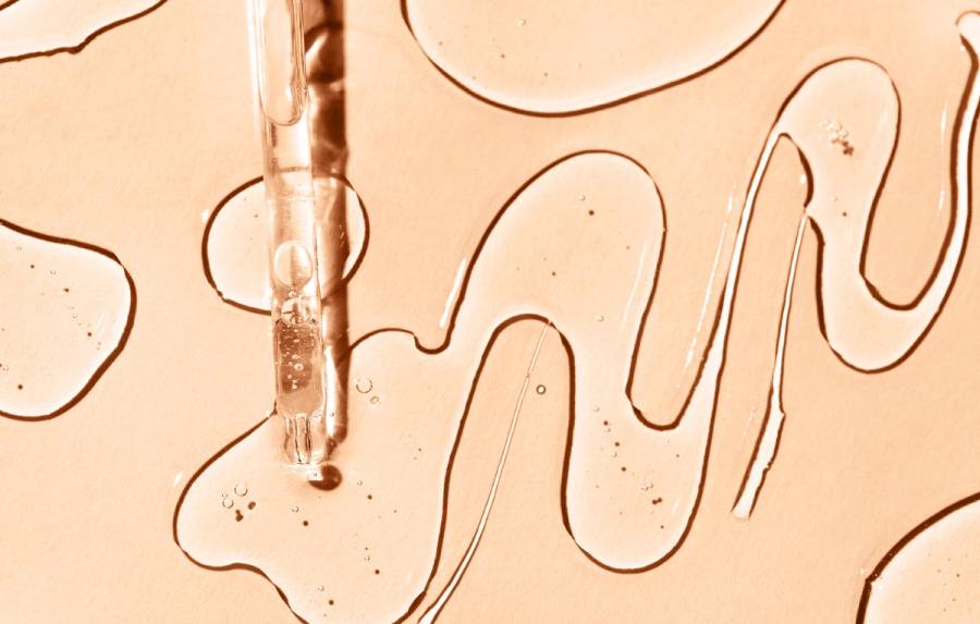 Close-up of a glass dropper releasing a serum, with squiggly lines of clear liquid on a peach-toned background