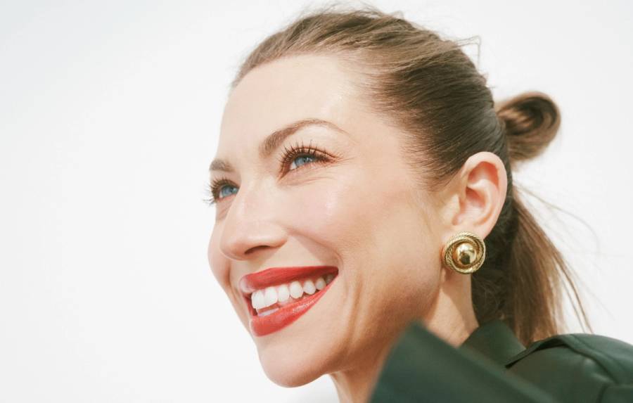 Stassi Schroeder smiles in a close-up portrait, wearing red lipstick, gold statement earrings and a dark green top with her hair pulled back.