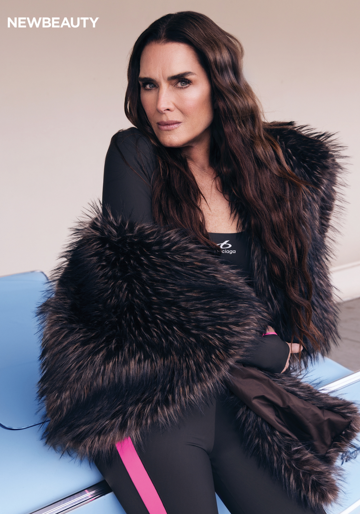 Brooke Shields sits on the hood of a vintage baby blue car wearing a dramatic fur stole over a Balenciaga sports top and black leggings with pink stripe detailing for NewBeauty's summer 2026 cover shoot