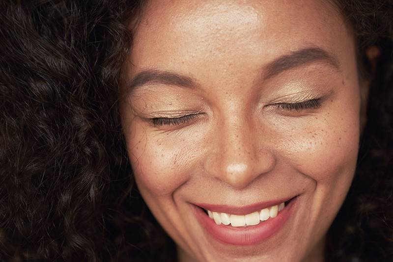 Closeup portrait of beautiful young woman with curly hair style and closed eyes. The woman has white toothy smile and freckles. The mulatto is a representative of a mixed race, an African American and a white race.