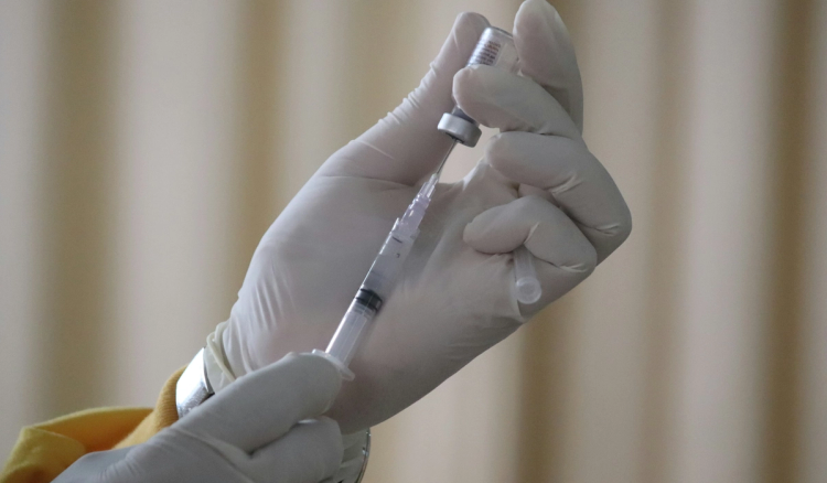A gloved hand in medical scrubs draws vaccine solution from a small glass vial into a syringe