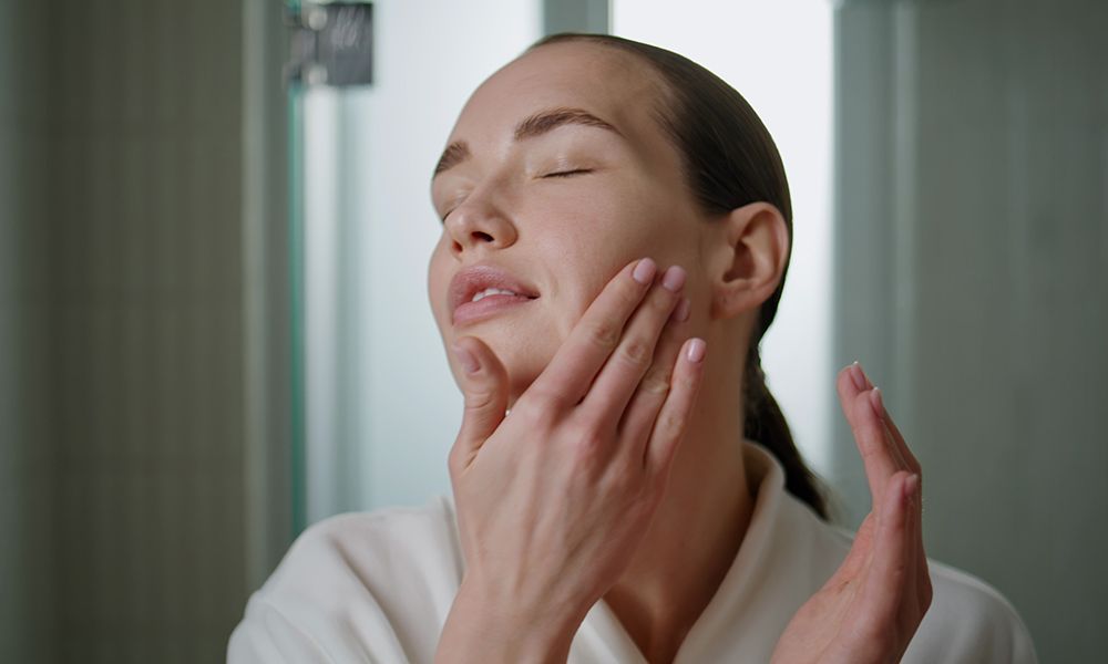 Beauty girl massaging serum on face closeup. Attractive woman applying moisturizing cream in daily routine at home. Serene young lady looking camera rubbing rejuvenating cosmetics in skin in bathroom.