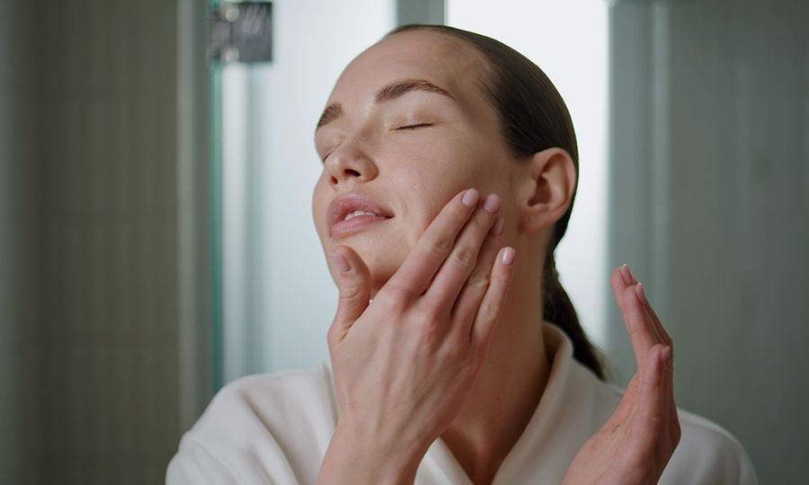 Beauty girl massaging serum on face closeup. Attractive woman applying moisturizing cream in daily routine at home. Serene young lady looking camera rubbing rejuvenating cosmetics in skin in bathroom.