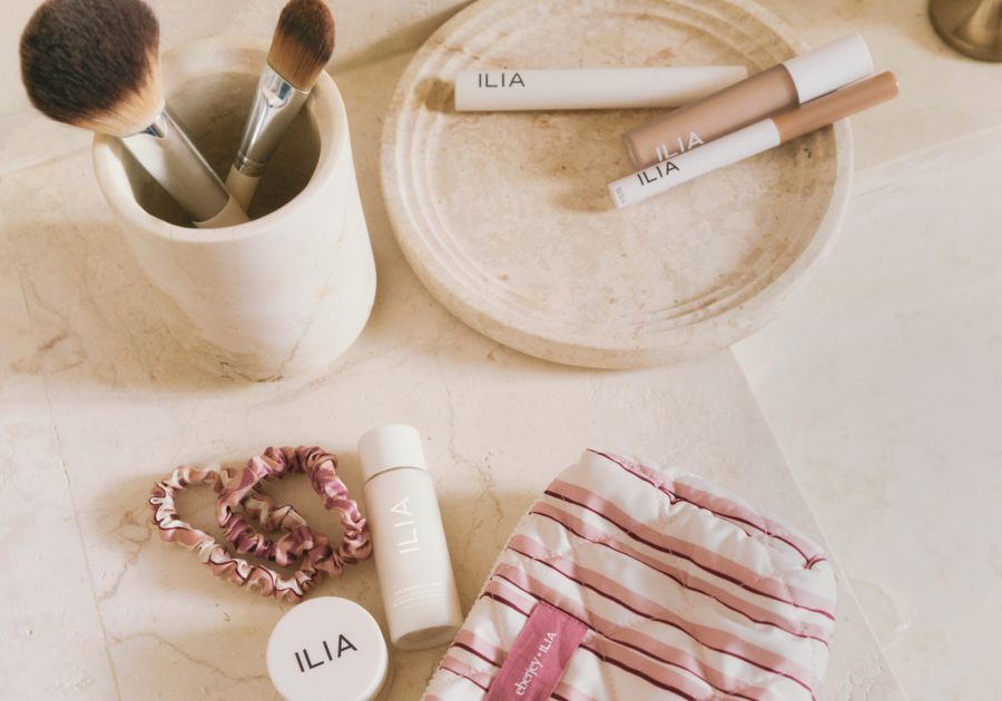 Flat lay of ILIA beauty products on a marble countertop, including makeup brushes in a ceramic cup, a concealer and lip product on a stone tray, a serum and balm, two pink scrunchies and a pink-and-white striped Eberjey pouch from the ILIA x Eberjey collaboration.