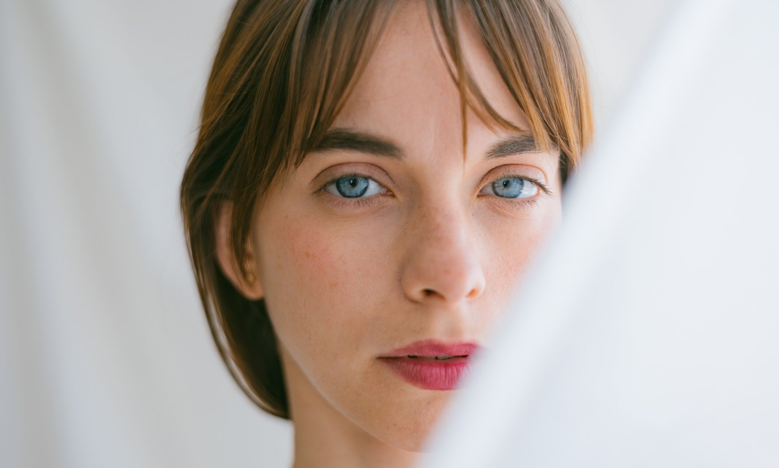 Studio portrait of a beautiful young woman with blue eyes looking at the camera with a serious face. With translucent curtains covering half of her face.