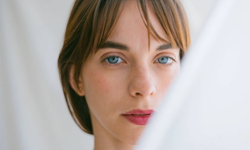 Studio portrait of a beautiful young woman with blue eyes looking at the camera with a serious face. With translucent curtains covering half of her face.