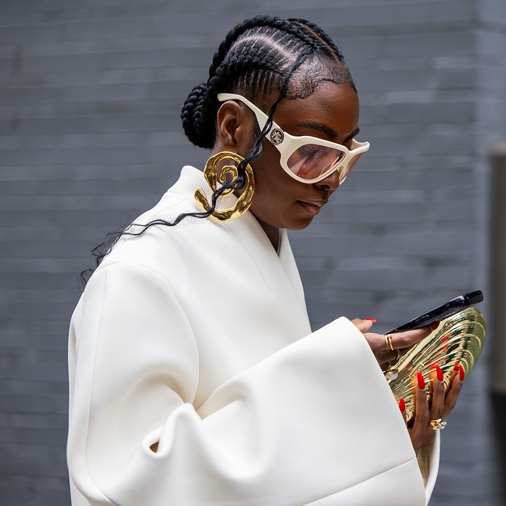 woman walking with braided hairstyle wearing a white coat and sunglasses looking at phone